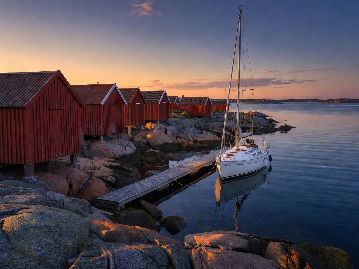 Red wooden boathouses on granite rocks in the Åland archipelago at dusk, with a small yacht moored to a jetty